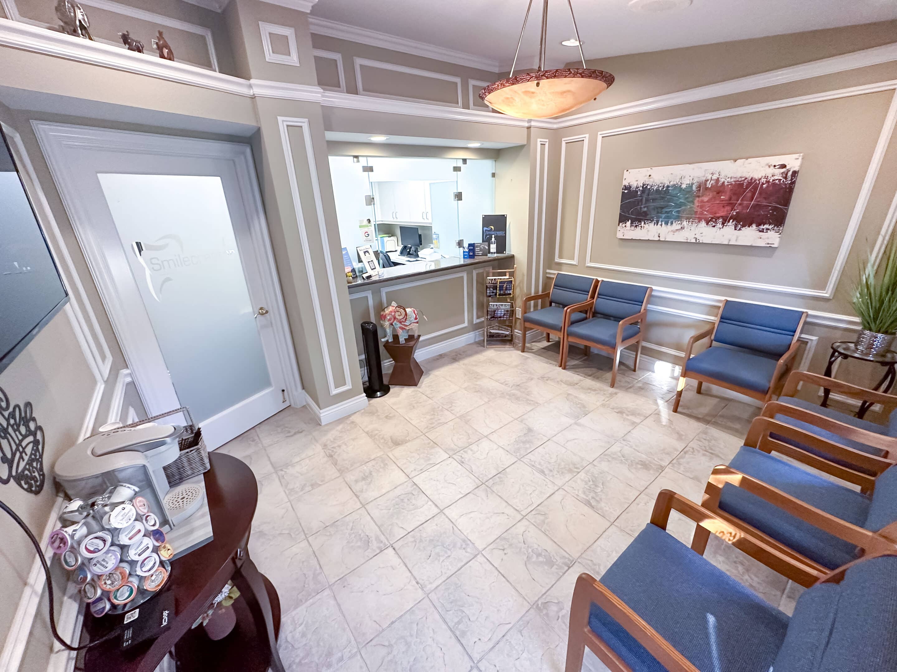A view of the waiting room at Smilecreator's Naples location, where various cushioned chairs are arranged and the front desk can be seen. There is a coffee maker on a table and the edge of a television screen visible.